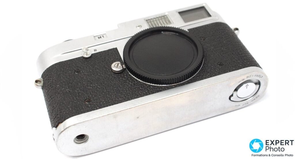 Leica M1 chromé vu de trois-quarts en plongée avec bouchon de monture noir, plaque M1 et vis R visibles.