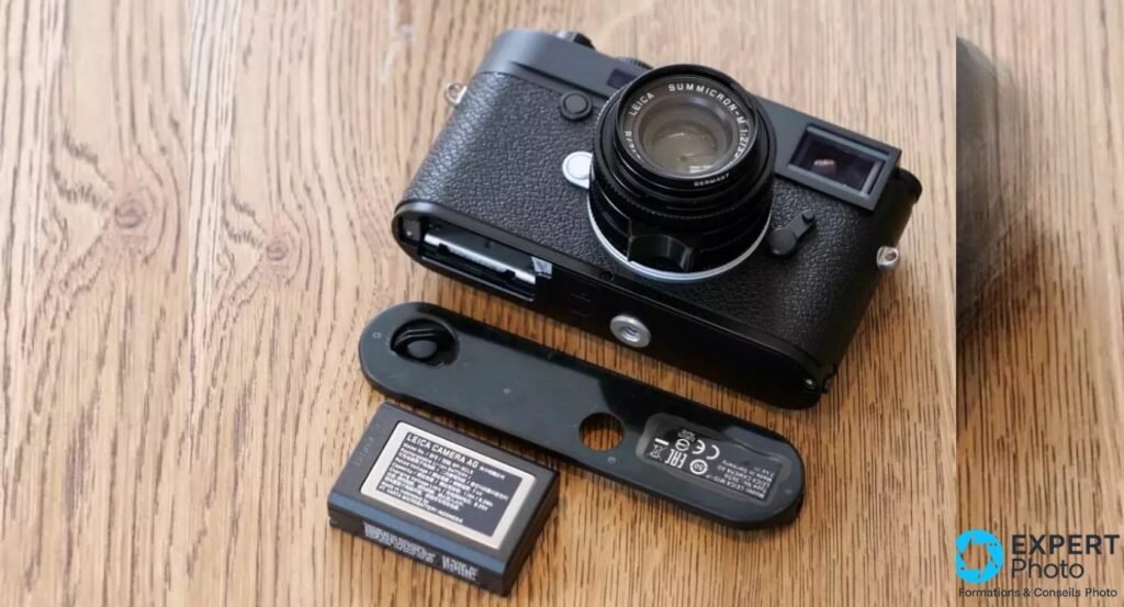 Leica M10-P noir, semelle ouverte posée à côté avec batterie BP-SCL5 et objectif Summicron-M monté, sur parquet chêne. 