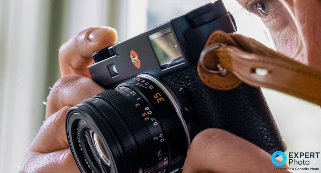  Leica M11 noir tenu à hauteur d'œil par un photographe, APO-Summicron-M 35 mm monté, dragonne en cuir tan visible.