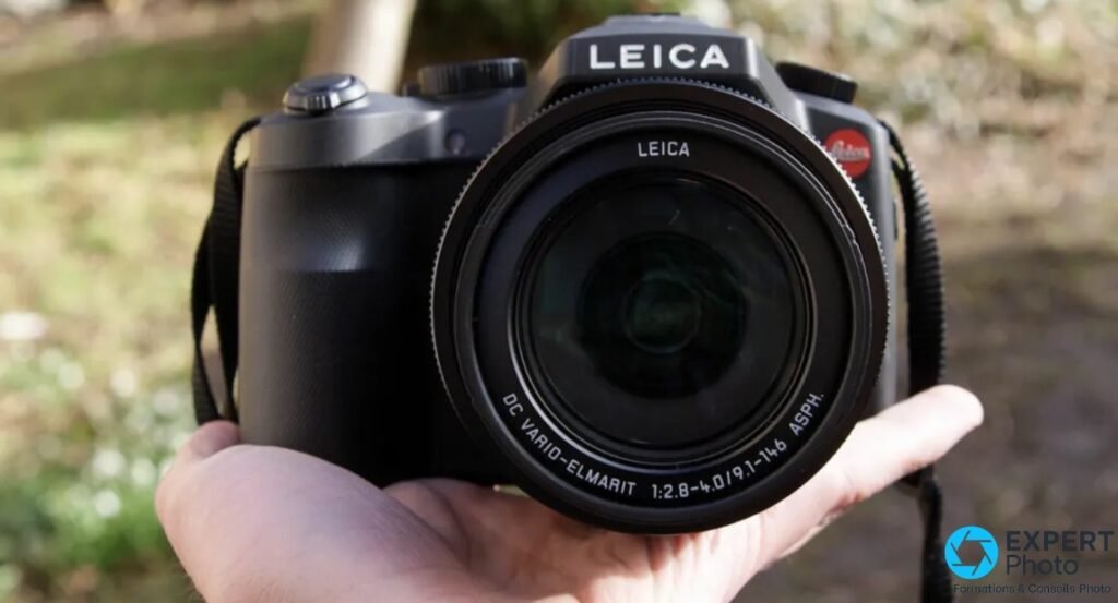 Leica V-Lux Typ 114 tenu à une main en extérieur, gabarit de bridge expert visible face à la paume, sangle noire 