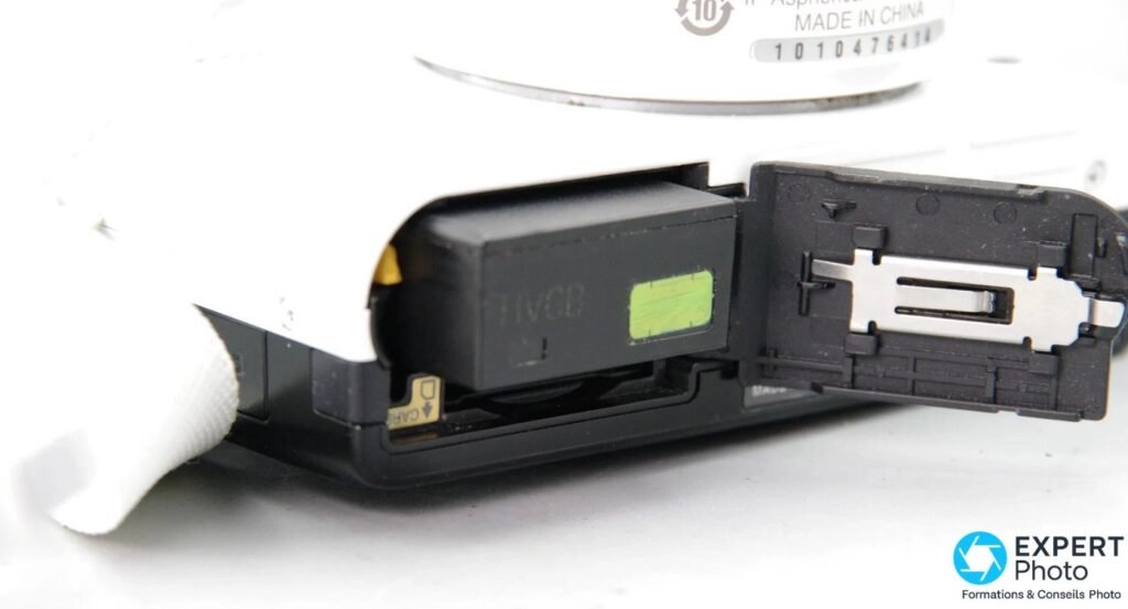 Trappe batterie et carte du Nikon 1 J2 ouverte laissant voir la batterie EN-EL20 insérée et le logement carte SD