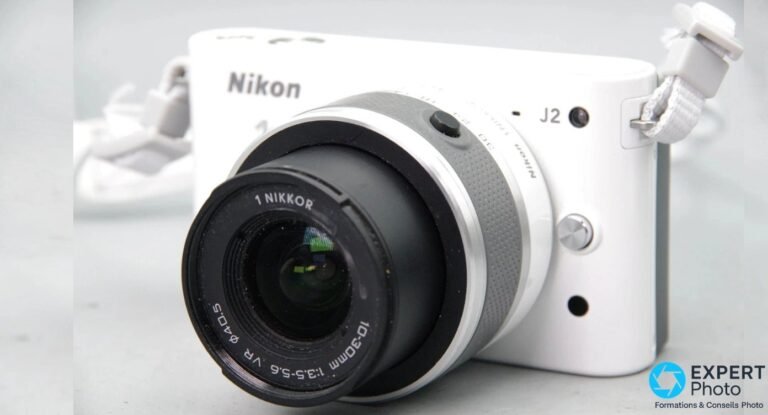 Nikon 1 J2 blanc posé sur fond neutre avec objectif 1 NIKKOR 10-30mm VR monté, dragonne blanche, vue de trois-quarts avant