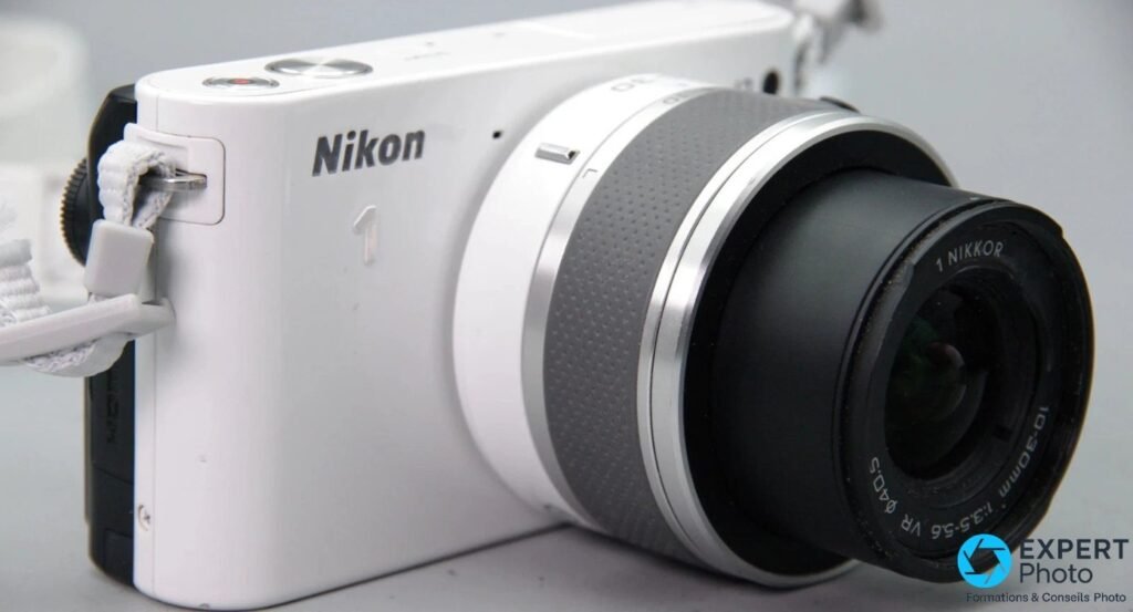 Nikon 1 J2 blanc vu de face légèrement en plongée, logo Nikon 1 visible, objectif 10-30mm VR monté, dragonne blanche 