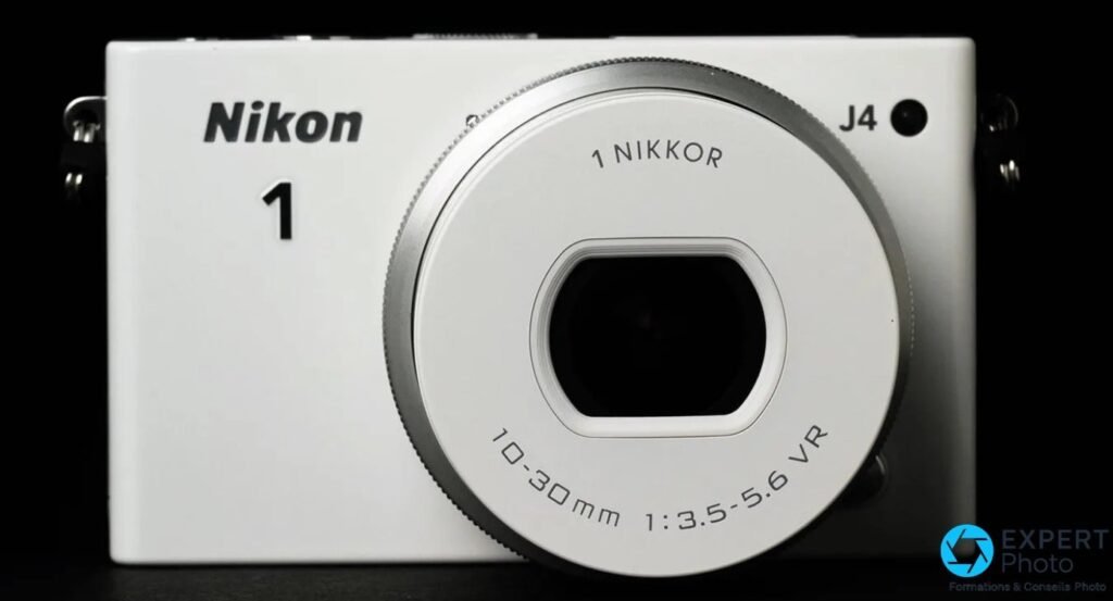 Nikon 1 J4 blanc vu de face avec objectif 1 NIKKOR 10-30mm f/3.5-5.6 VR monté et bouchon en place sur fond noir