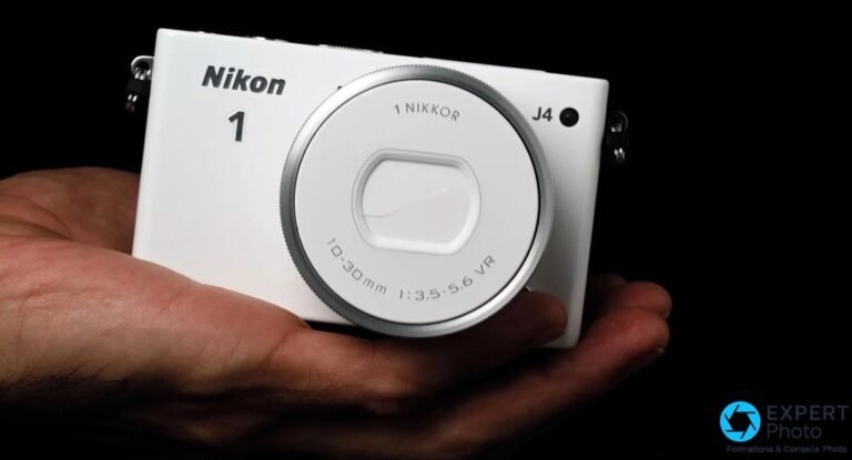 Nikon 1 J4 blanc posé dans la paume d'une main d'homme sur fond noir, objectif 10-30mm rétracté bouchon en place
