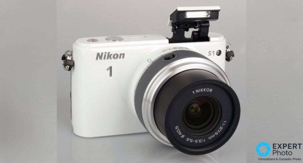 Nikon 1 S1 blanc en trois-quarts avant, flash intégré pop-up déployé au-dessus du boîtier, objectif de kit 11-27,5 mm monté.
