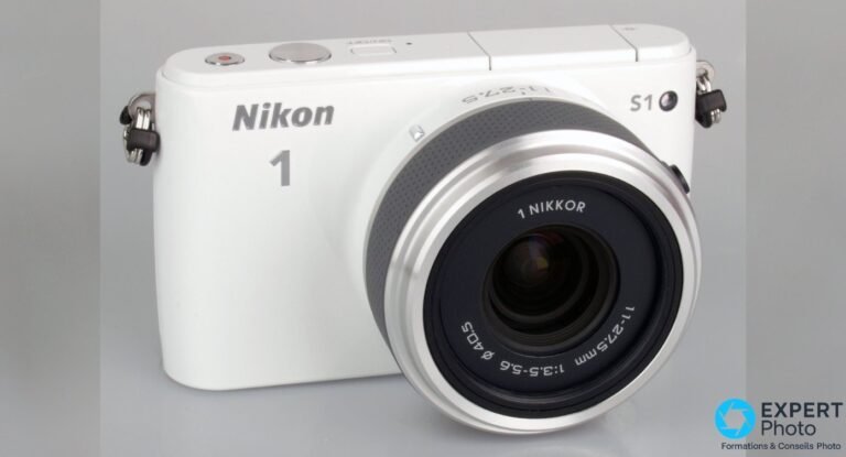Hybride Nikon 1 S1 blanc en trois-quarts avant, objectif 1 Nikkor 11-27,5 mm monté sans capuchon, fond gris uni propre.