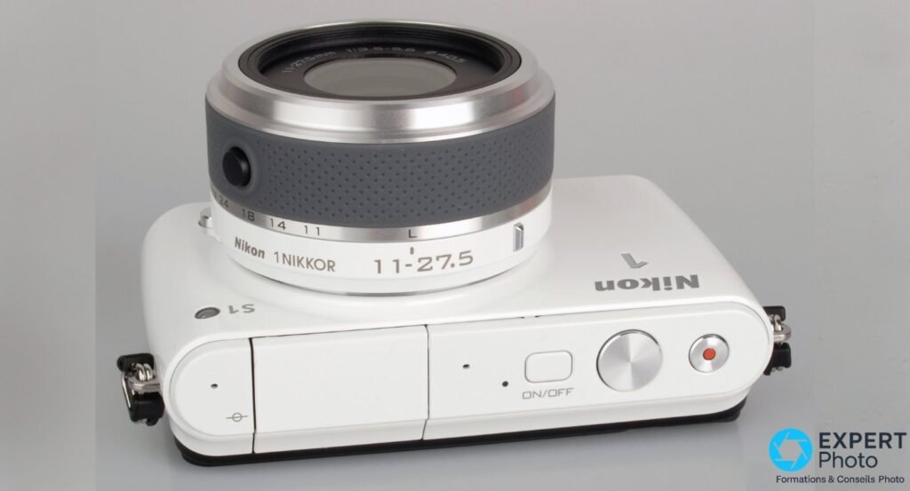 Dessus du Nikon 1 S1 blanc, objectif 11-27,5 mm replié, déclencheur argenté, bouton ON/OFF et touche vidéo rouge visibles.