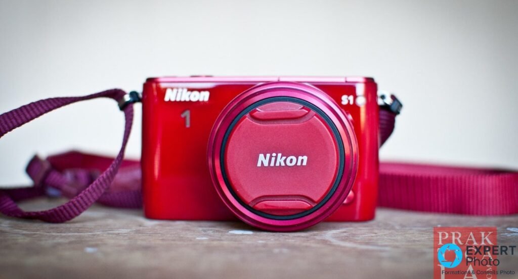 Boîtier hybride Nikon 1 S1 rouge de face sur plan de travail bois, capuchon Nikon rouge fixé, sangle bordeaux enroulée.