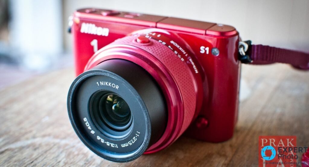 Nikon 1 S1 rouge de face avec objectif 1 Nikkor VR 11-27,5 mm f/3.5-5.6 déployé sans capuchon, posé sur plan de travail bois. 
