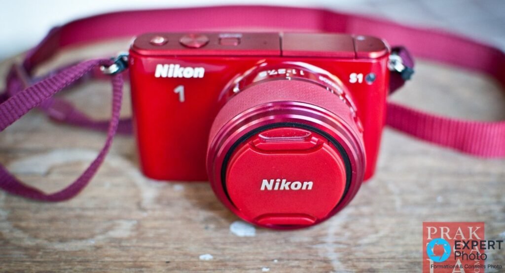 Nikon 1 S1 rouge en trois-quarts avant avec objectif 1 Nikkor 11-27,5 mm rétracté et capuchon rouge, sangle bordeaux visible.