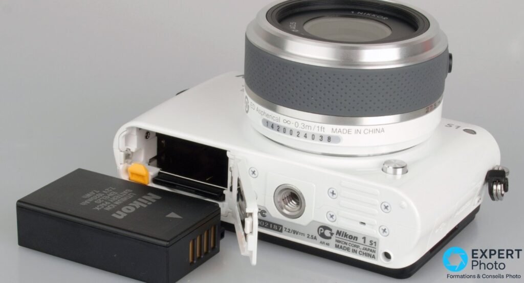 Dessous du Nikon 1 S1 blanc, trappe batterie ouverte, batterie Nikon EN-EL20 posée à côté, filetage trépied visible.