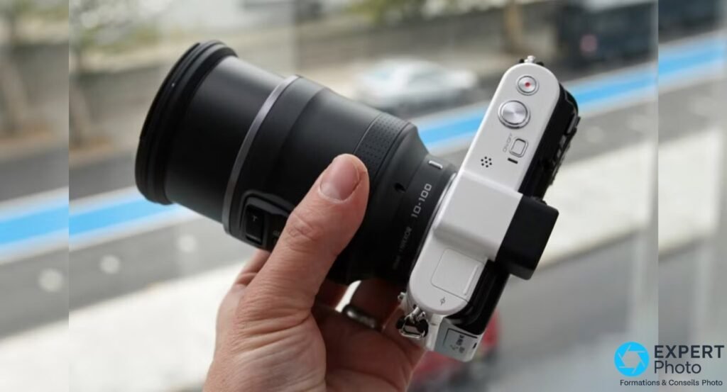 Nikon 1 V1 blanc tenu en main, équipé du 1 NIKKOR VR 10-100mm téléobjectif, déséquilibre visible