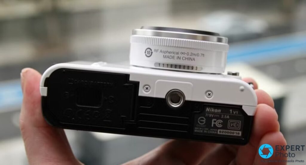 Dessous du Nikon 1 V1 blanc : trappe batterie, filetage trépied, étiquette Nikon 1 V1 et numéro de série 