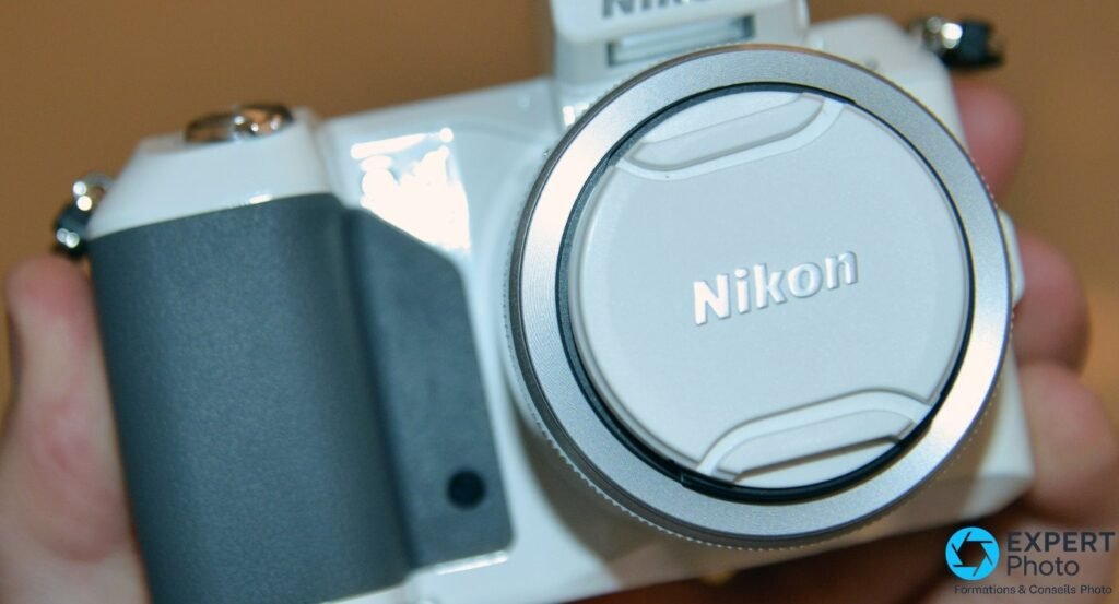 Nikon 1 V2 blanc tenu à deux mains, vue avant 3/4, objectif blanc avec bouchon Nikon au premier plan, flash intégré en haut