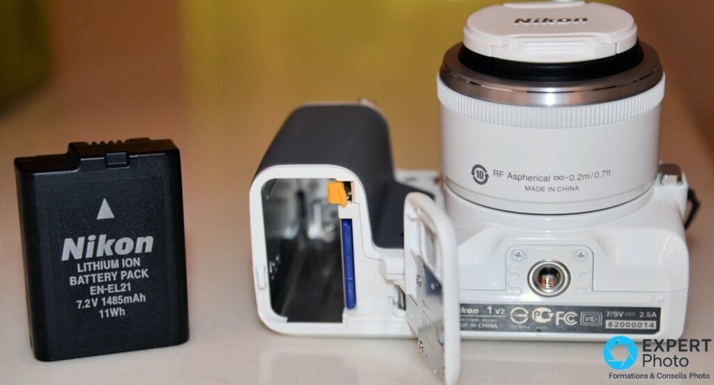 Nikon 1 V2 blanc objectif blanc monté, trappe batterie et carte SD ouverte sur le côté, batterie Nikon EN-EL21 à gauche 