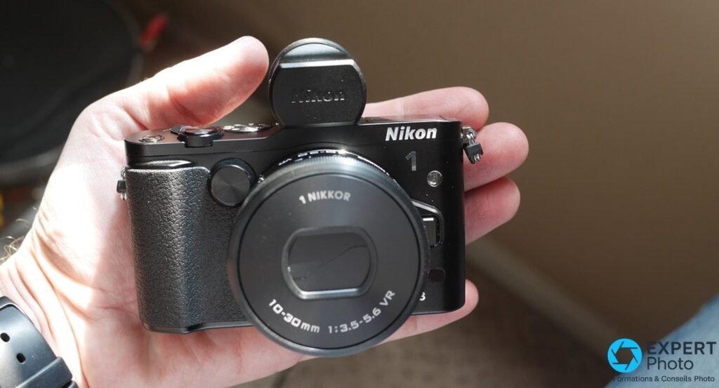Nikon 1 V3 noir tenu dans la main avec objectif 1 NIKKOR 10-30 mm VR, bouchon rond Nikon posé au-dessus
