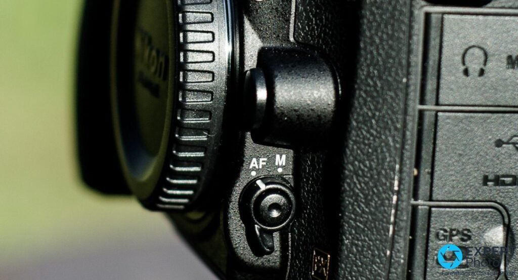 Gros plan sélecteur AF/M du Nikon D600 sur la tranche gauche, bague de zoom objectif et prises latérales visibles.