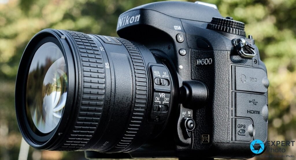 Nikon D600 vu en trois-quarts gauche avec zoom VR, panneaux latéraux fermés HDMI, USB, MIC et GPS visibles.