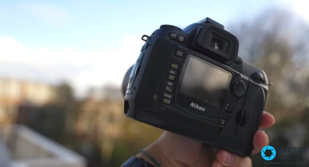 Nikon D70s tenu en main, face arrière, écran LCD 2 pouces et boutons MENU ISO WB QUAL visibles, ciel nuageux en fond. 