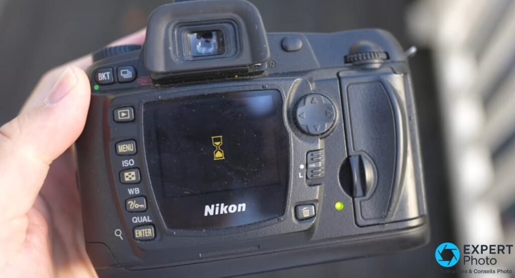 Nikon D70s face arrière en main, écran LCD avec icône sablier de buffer, boutons MENU ISO WB QUAL ENTER et viseur optique.