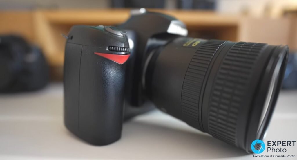 Nikon D70s posé sur surface, vue avant droite, objectif zoom grand diamètre monté, grip et flash fermé en premier plan.