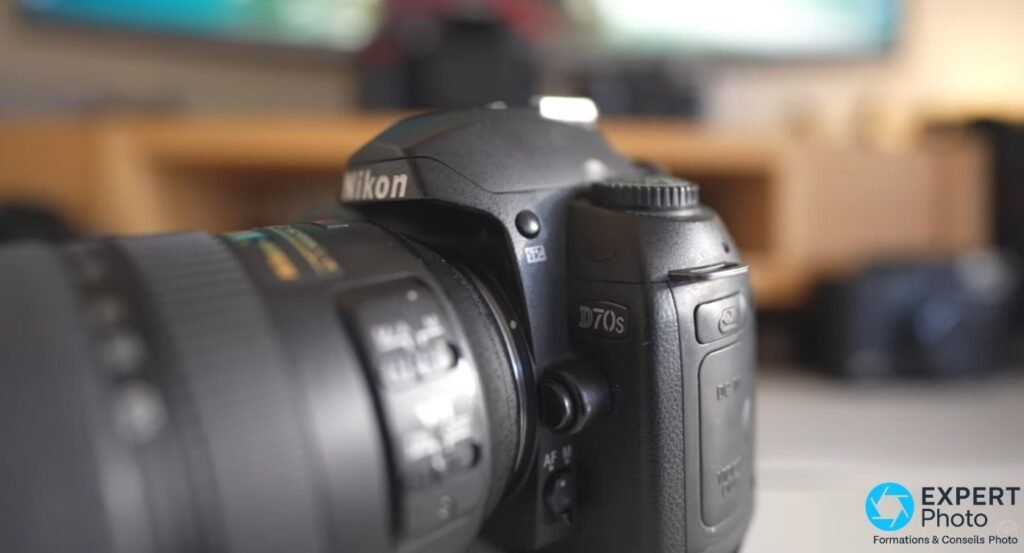 Nikon D70s posé sur table, vue avant latérale, inscription D70s sur le boîtier et objectif zoom monté en lumière intérieure. 