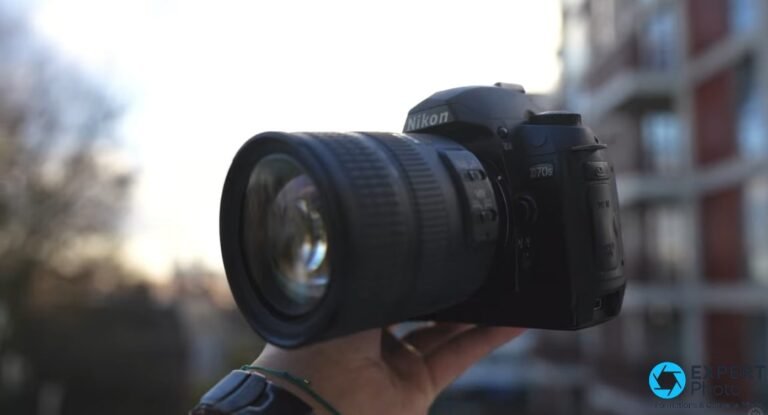 Nikon D70s tenu en main en extérieur, vue avant trois quarts, label D70s lisible, objectif zoom monté, bâtiment en fond.