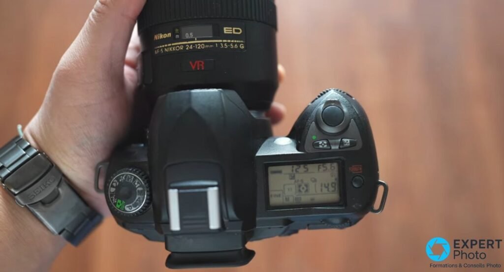 Nikon D70s vu de dessus tenu en main, écran LCD supérieur affichant 12.5 et F5.6, Nikon AF-S 24-120mm monté.