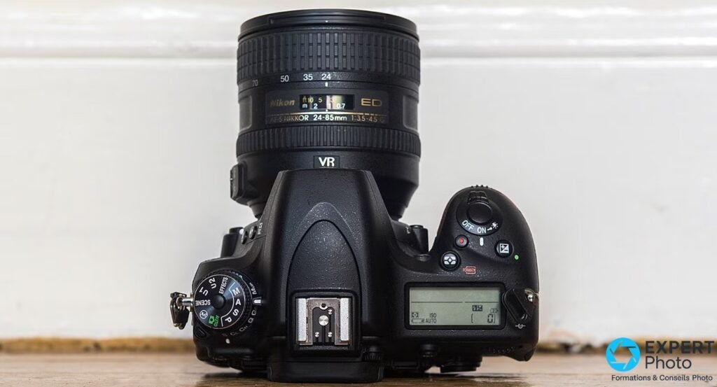 Nikon D750 vu de dessus, molette de modes P A S M visible, objectif Nikon 24-85mm monté, écran LCD supérieur allumé