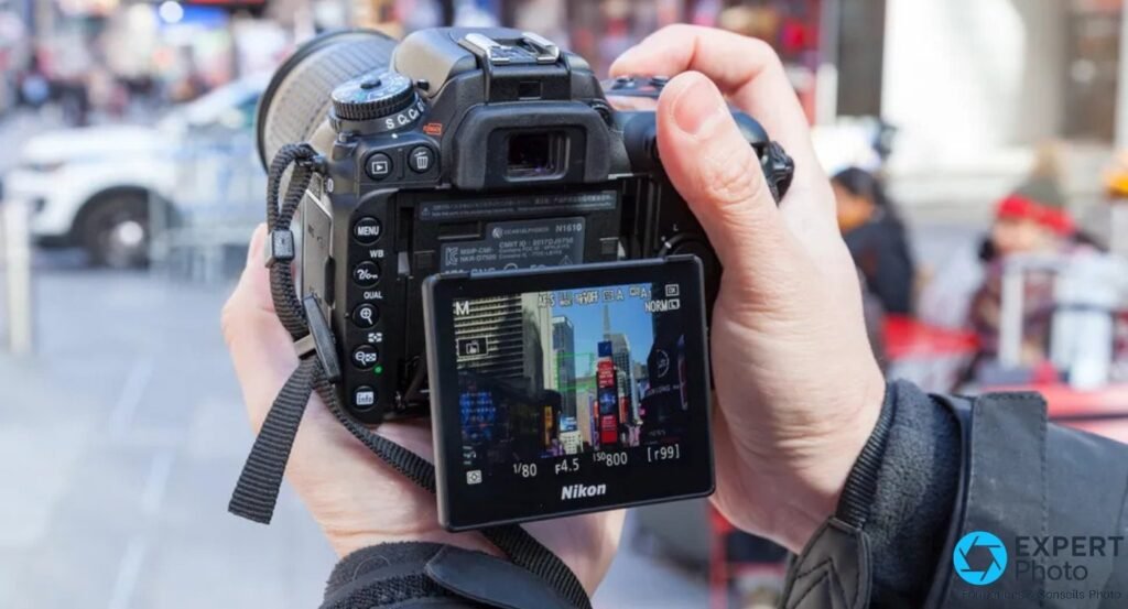 Nikon D7500 tenu à deux mains, écran inclinable en LiveView sur scène urbaine, mode M, 1/80s, f4.5, ISO 800 affichés