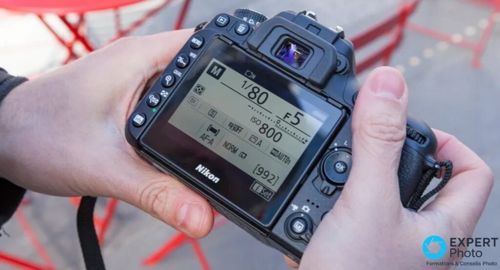 Dos du Nikon D7500 tenu à deux mains, LCD mode M, 1/80s, F5, ISO 800, AF-A, 992 vues restantes, fond extérieur rouge