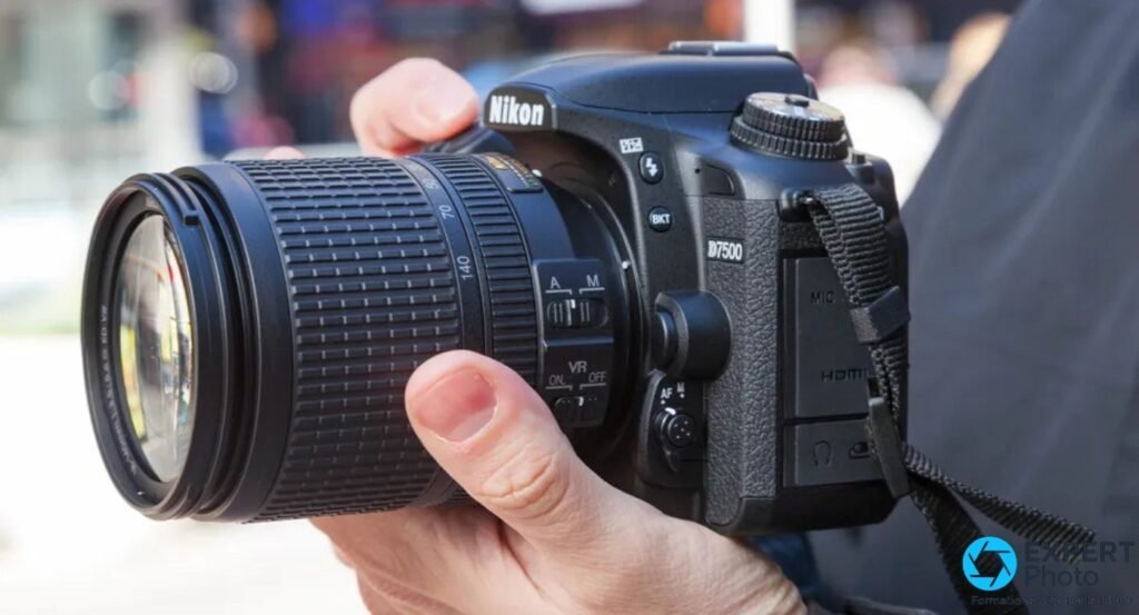 Main droite tenant un Nikon D7500 avec objectif zoom 18-140mm VR, bague de zoom et commutateur VR visibles, fond urbain flou