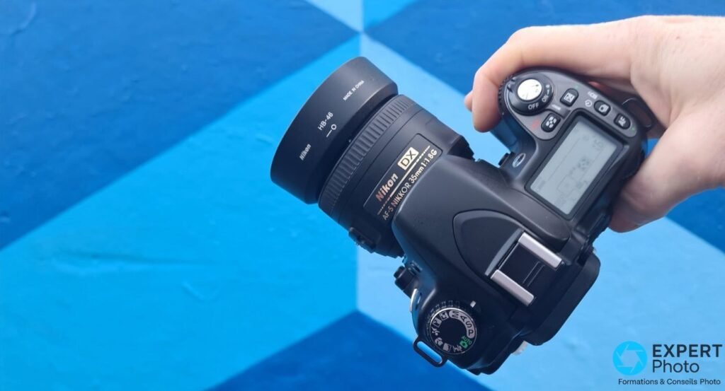 Main tenant un Nikon D80 par le dessus avec objectif Nikkor DX 35mm f/1.8G et pare-soleil HB-46, molette de modes visible 