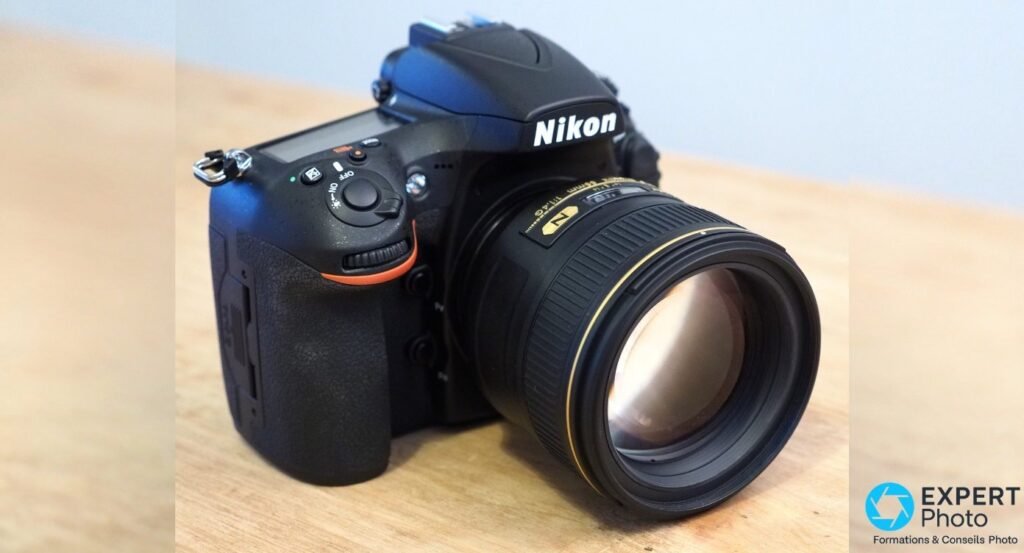 Nikon D810A en vue trois quarts avant plongeante, objectif Nikon 85mm monté, accent orange sur grip, logo lisible