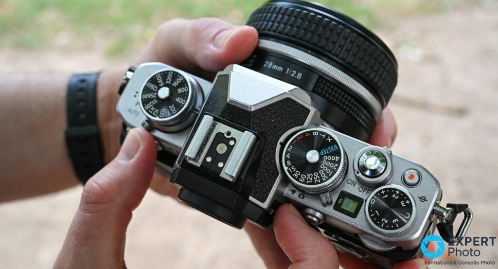 Nikon Z fc tenu à deux mains, vue trois quarts dessus extérieur, molettes physiques et objectif 28 mm f/2.8 visibles.