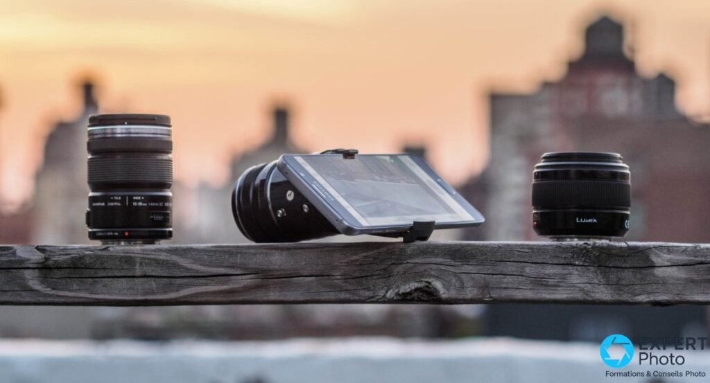 Olympus AIR A01 avec smartphone Samsung clipsé, entouré d'objectifs Micro Four Thirds sur traverse bois, coucher de soleil