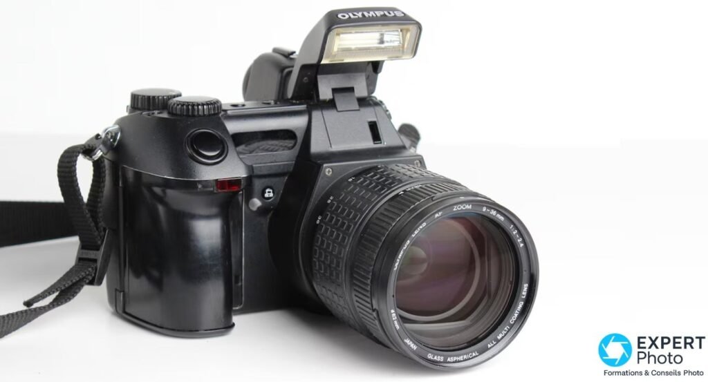 Olympus E-10 en 3/4 avant avec flash intégré déployé marqué OLYMPUS et objectif zoom 9-36 mm avec pare-soleil monté. 