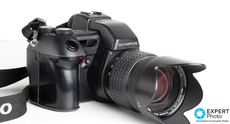 Olympus E-10 en 3/4 profil droit, logo OLYMPUS sur le prisme, objectif zoom 9-36 mm avec pare-soleil pétale noir monté.