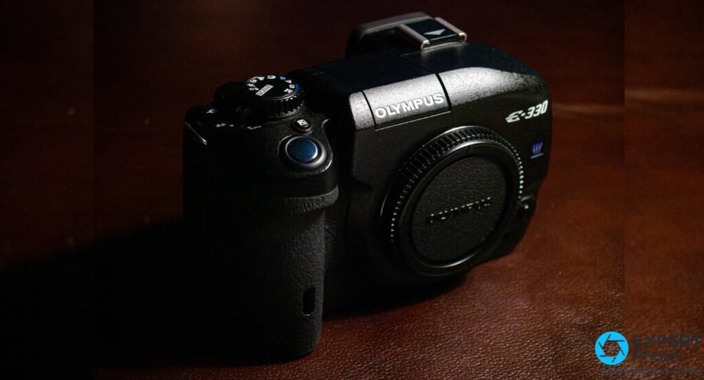 Olympus E-330 noir vu de 3/4 avant sur fond sombre, bouchon de monture Olympus en place, molette des modes visible 