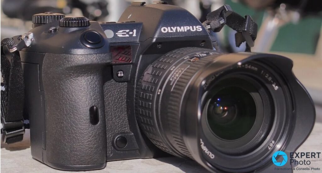 Olympus E-1 avec objectif Zuiko Digital 14-54mm f/2.8-3.5 et pare-soleil monté, vue 3/4 avant en extérieur. 