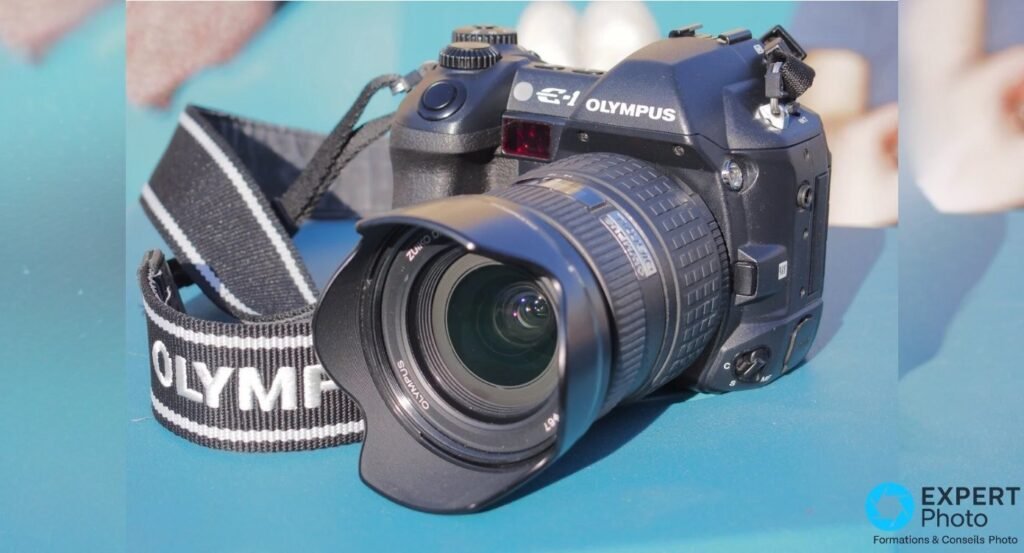 Olympus E-1 avec zoom Zuiko Digital 14-54mm et pare-soleil, sangle Olympus au premier plan, fond turquoise.