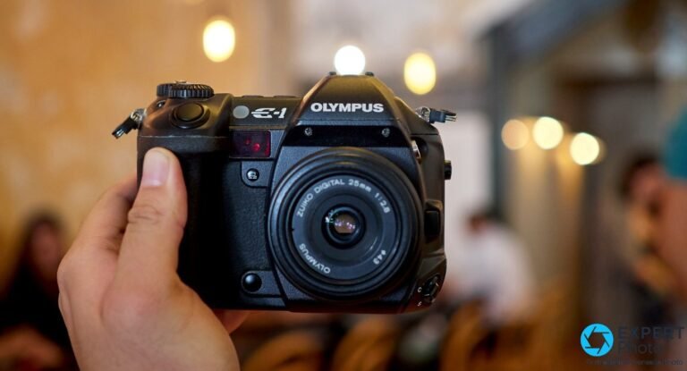 Olympus E-1 avec pancake Zuiko Digital 25mm f/2.8 tenu en main, ambiance intérieure avec bokeh de lampes chaudes.