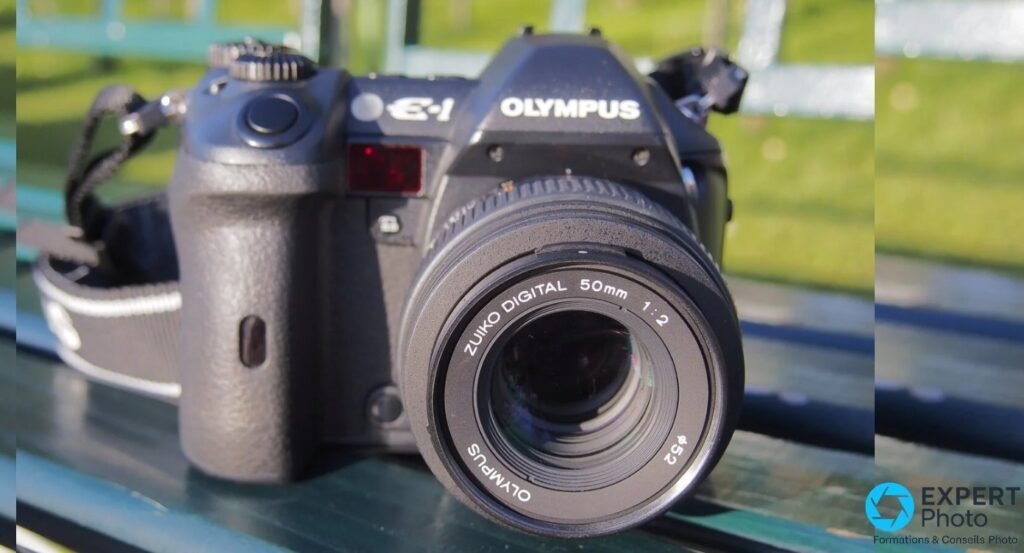 Olympus E-1 équipé du Zuiko Digital 50mm f/2 Macro, vue de face sur banc vert avec pelouse en arrière-plan. 