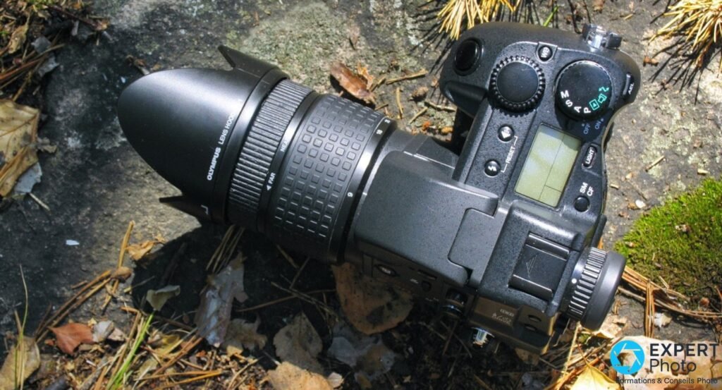 Olympus E-20 vu de dessus posé sur sol forestier, objectif avec pare-soleil, écran LCD et molette de modes visibles