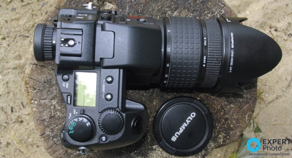 Olympus E-20 vu de dessus avec objectif zoom 9-36 mm, focales gravées, pare-soleil Olympus LH-1 et bouchon à côté