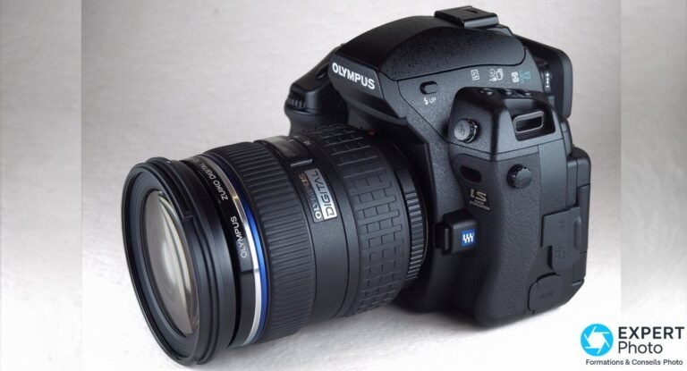 Olympus E-3 noir en vue trois quarts avant, objectif Zuiko Digital à bague bleue monté, sur fond studio gris.