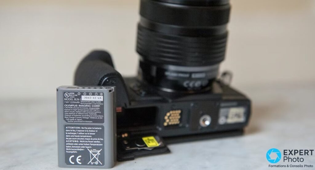 Olympus OM-D E-M1 avec batterie BLN-1 sortie et trappe ouverte, carte SD insérée et objectif monté en arrière-plan.