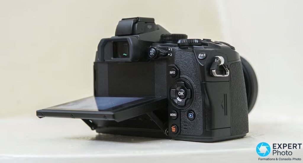 Olympus OM-D E-M1 de profil gauche, écran tactile inclinable déployé vers le bas, boutons AEL/AFL et Fn1 visibles. 