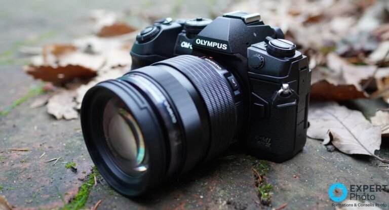 Olympus OM-D E-M1 Mark III équipé d'un zoom M.Zuiko PRO avec bouton L-Fn, posé sur feuilles mortes, vue 3/4 avant.